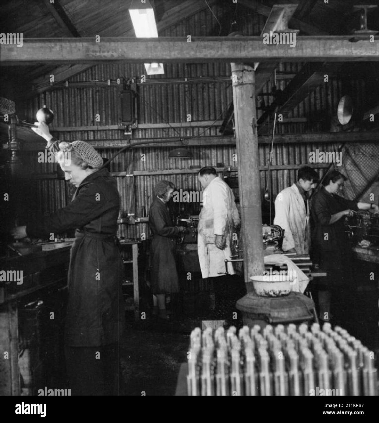 Sten Gun Production in Britain, 1943 Mrs Lushby (left) and colleagues at work in a small 'factory' producing breech blocks from steel bars. This 'factory', previously used as a hen house, was owned by Mr C W (Charles) Packard, an engineer and former haulage contractor, and was probably in Welwyn Garden City. In the foreground, rows of finished 'block pieces' can be seen, ready for the next stage of production. Stock Photo