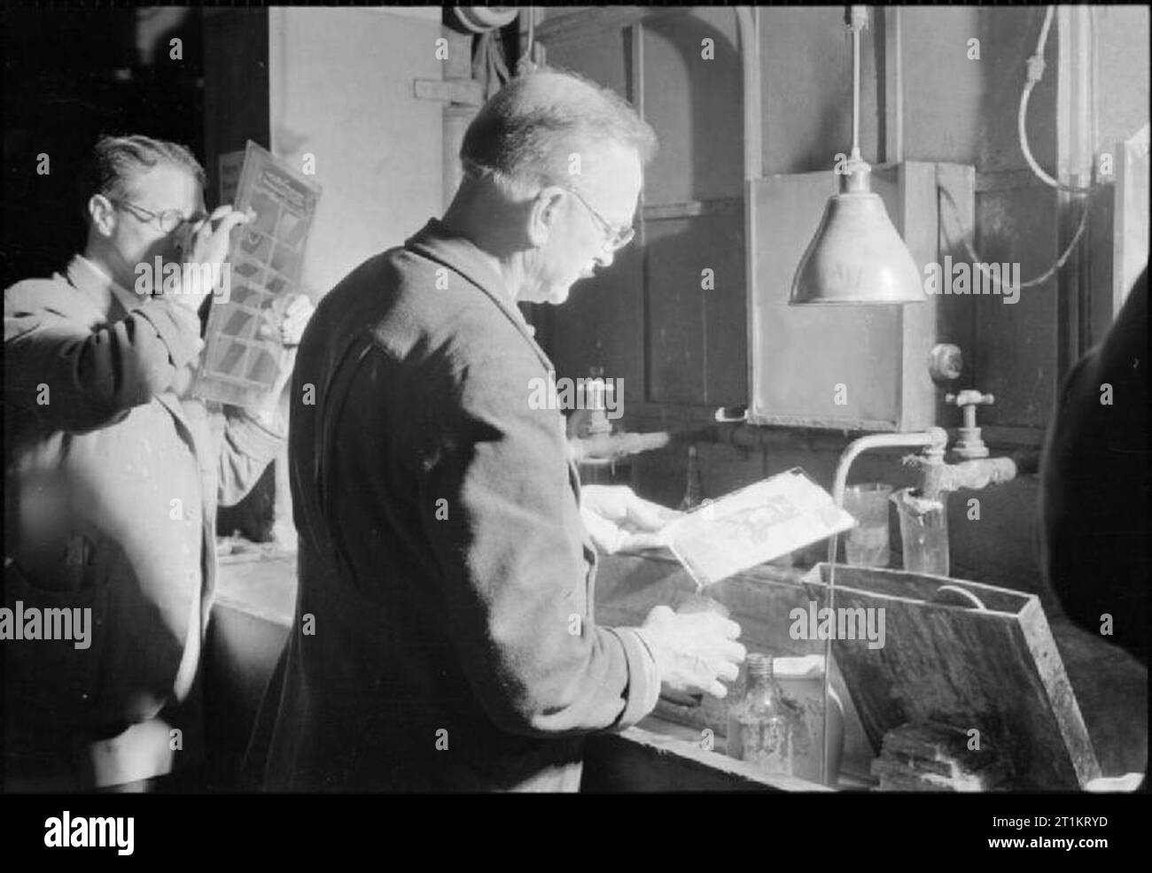 The Makings of a Modern Newspaper- the Production of 'the Daily Mail' in Wartime, London, UK, 1944 In the photographic darkrooms at the Daily Mail, two photographers are at work. One is examining a sheet of negatives with a magnifier, and the second is rinsing a plate before making a half-tone block. The image will be etched onto a copper plate before being made up 'on the stone' as part of the newspaper page. Stock Photo