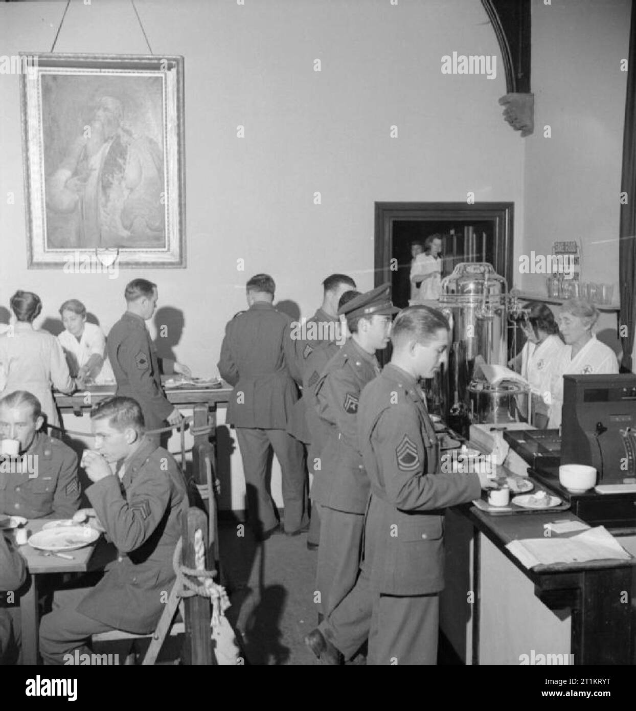 American Red Cross Service Club, Norwich- Life at the Club at the Bishop's Palace, Norfolk, England, UK, 1943 American service personnel queue along the counter of the buffet which lines the walls of the cafeteria at the American Red Cross Service Club. The room was previously the dining room in the Bishop's Palace. British staff, both paid and volunteer, serve meals to the men. According to the original caption, hot food, sandwiches, tea, coffee and coca-cola is always available, day and night, and Bishop Herbert calls into the cafeteria every day for his morning coffee. Stock Photo