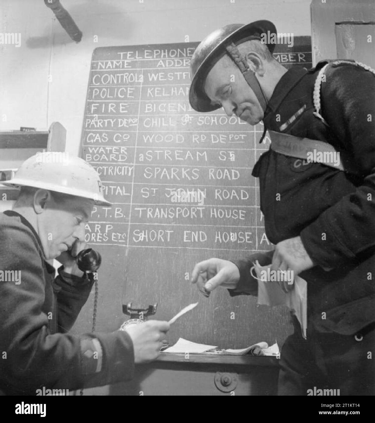 The Reconstruction of 'an Incident'- Civil Defence Training in Fulham, London, 1942 An Air Raid Warden gives his report of the local incident to a colleague at the ARP post, which is then telephoned through to the Control Centre. Behind them, a blackboard lists the names and addresses of the various services to contact, including the electricity company, the water board and the fire station. The names of the streets listed are false, and just serve as an illustration during this reconstruction. Stock Photo