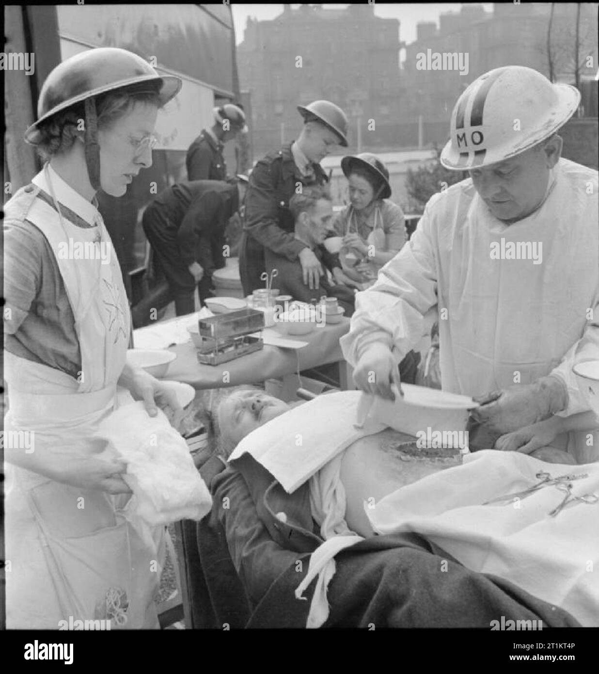 The Reconstruction of 'an Incident'- Civil Defence Training in Fulham, London, 1942 The Medical Officer of the Mobile Medical Unit treats a casualty with a severe abdominal wound, before he is transferred to hospital. The Medical Officer (with the letters 'MO' on his steel helmet) is assisted by a nurse. In the background, Civil Defence Wardens help another casualty to drink a cup of tea. This photograph was taken at the end of Edith Villas, where it butts up to West Cromwell Road/Conan Street, looking towards Conan Street, which runs at right angles to Edith Villas. Stock Photo