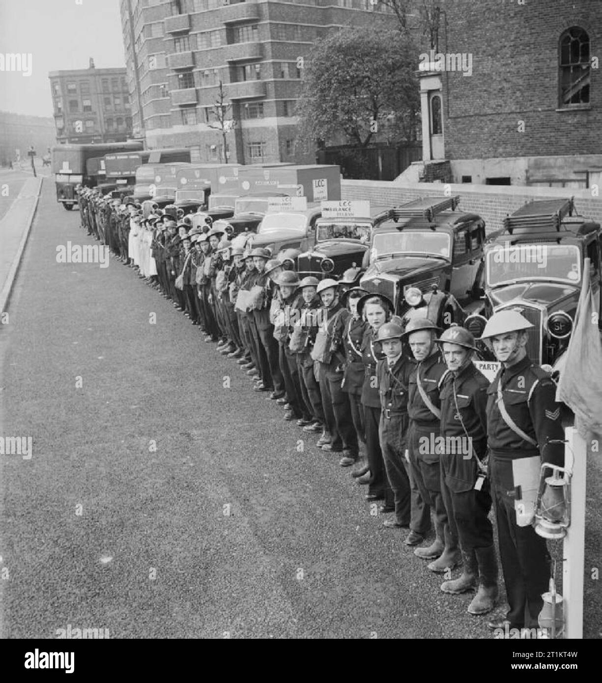 The Reconstruction of 'an Incident'- Civil Defence Training in Fulham, London, 1942 Members of various Civil Defence services, including ambulance workers, rescue and stretcher parties, Civil Defence wardens and nursing staff line up with their vehicles at the end of a large-scale Civil Defence training exercise in Fulham. Twelve vehicles and fifty personnel, not including the Fire Service, were involved in this exercise. The Incident Officer, wearing a blue helmet, stands next to his blue lamp and flag in the foreground. This photograph was taken on Conan Street, now West Cromwell Road, looki Stock Photo