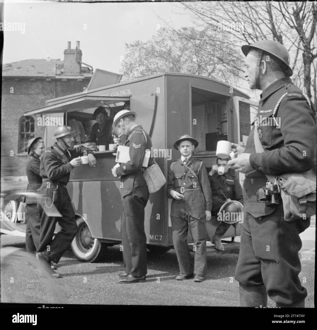 The Reconstruction of 'an Incident'- Civil Defence Training in Fulham, London, 1942 Civil Defence workers enjoy tea and refreshments at the mobile canteen during a break, now that the incident has been brought under control. As well as Civil Defence Wardens, also visible are a boy messenger and the Incident Officer, wearing a blue helmet, who can been seen lighting a cigarette in the centre of the photograph. A female Civil Defence Warden waits her turn at the counter. This photograph was almost certainly taken on Conan Street/West Cromwell Road (see also D 7916 and D 7917), near to where it j Stock Photo