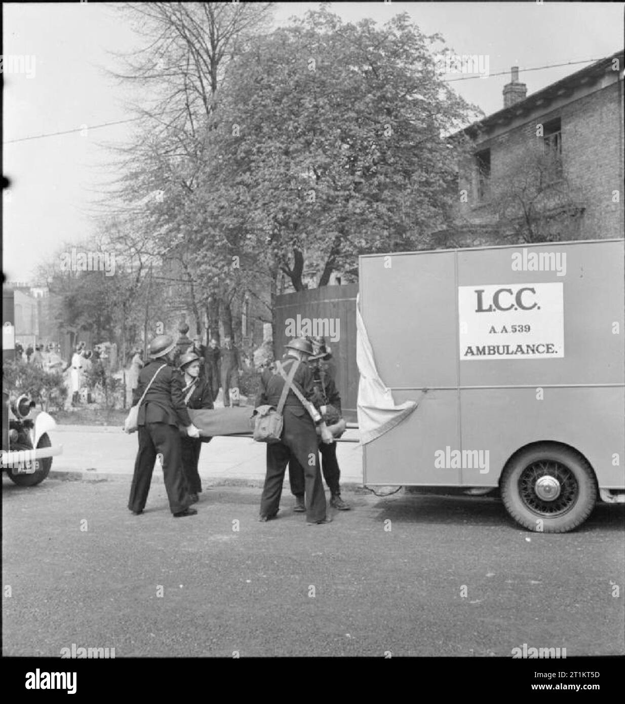 The Reconstruction of 'an Incident'- Civil Defence Training in Fulham, London, 1942 Female Ambulance drivers help men of the 'light rescue' stretcher party to load a stretcher case into an ambulance. A walking wounded case can be seen in the background as he is guided to an ambulance car parked further up the street. This photograph was taken on West Cromwell Road/Conan Street, looking back down Edith Villas. The Mobile Medical Unit is just visible to the left of the photograph. Stock Photo