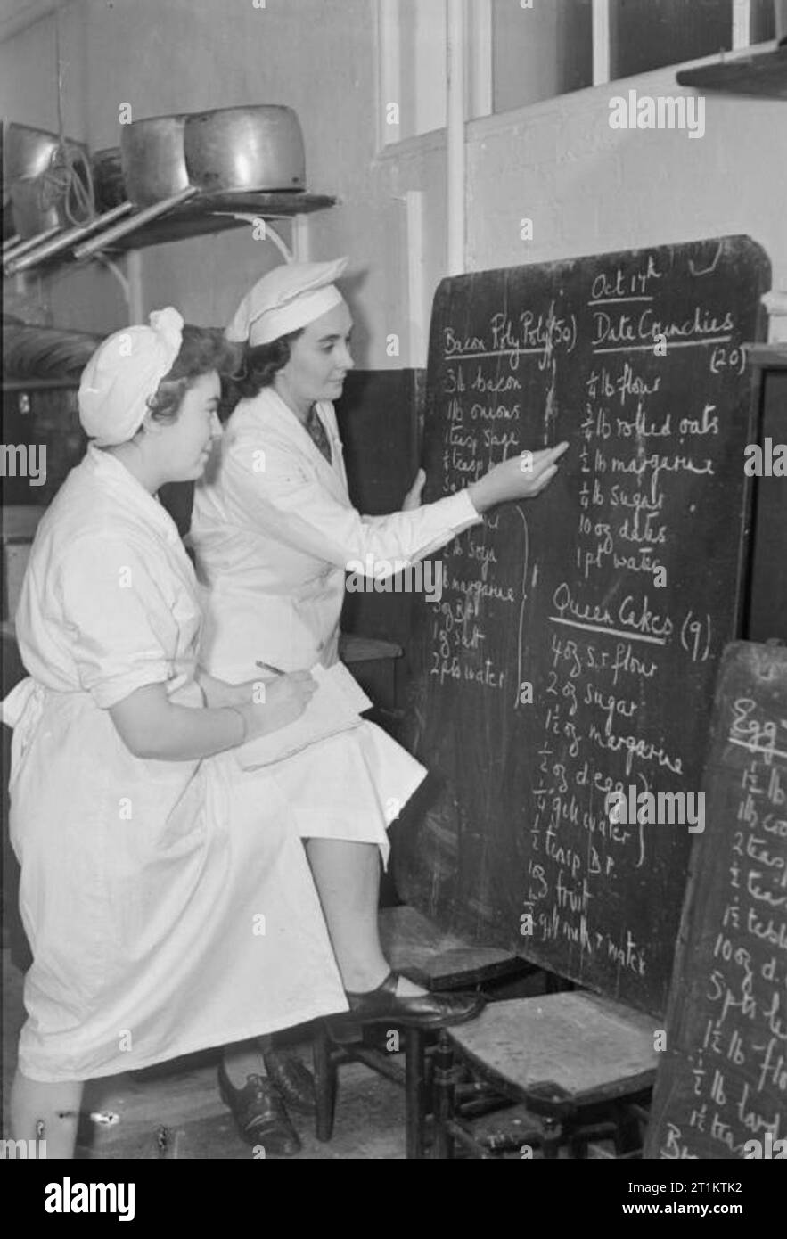 They Train To Be Cooks- Cookery Classes at the National Training College of Domestic Science, Westminster, London, England, UK, 1944 Upon arrival at the National Training College of Domestic Science at 8:30am, a student (left) takes down the day's menu from the blackboard in her notebook, as the instructor points to a particular ingredient. The recipes are for bacon roly poly (to serve 50), date crunchies (to serve 20) and queen cakes (to make 9). Stock Photo