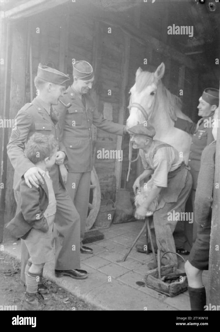 US Troops in An English Village- Everyday Life With the Americans in Burton Bradstock, Dorset, England, UK, 1944 At the blacksmith's shop in Burton Bradstock, Dorset, Sergeant R J Baier (left, smoking pipe, of 125 Park Avenue, Canandaigna, New York), Corporal David Roberts (of RR4 Albia, Iowa), more of their American GI colleagues and a local boy look on as blacksmith R Burton finishes shoeing a horse. According to the original caption, 'they have come to beg horseshoes for a game at the camp'. Stock Photo
