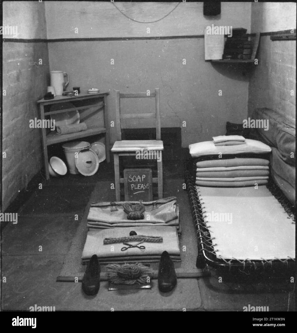 Wakefield Training Prison and Camp- Everyday Life in a British Prison, Wakefield, Yorkshire, England, 1944 A view of an inmate's room at Wakefield Prison. Clearly visible are the bed, a chair, several small shelves, and slop bucket. The rest of the inmate's belongings, such as a pair of shoes and a comb, have been set out neatly, ready for inspection. Chalked on a small blackboard are the words 'soap please'. Stock Photo