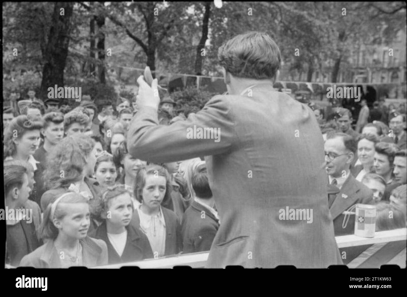 War Fair- Holidays at Home at a Fete in Russell Square, London, 1943 Film and stage actor Derek de Marnay stands at a microphone and holds aloft the very small, very green, banana being auctioned as part of the festivities in Russell Square. Many of the children pictured here will not have seen a banana before, due to the scarcity of this fruit in wartime. According to the original caption, the banana fetched ?GBP5. Stock Photo