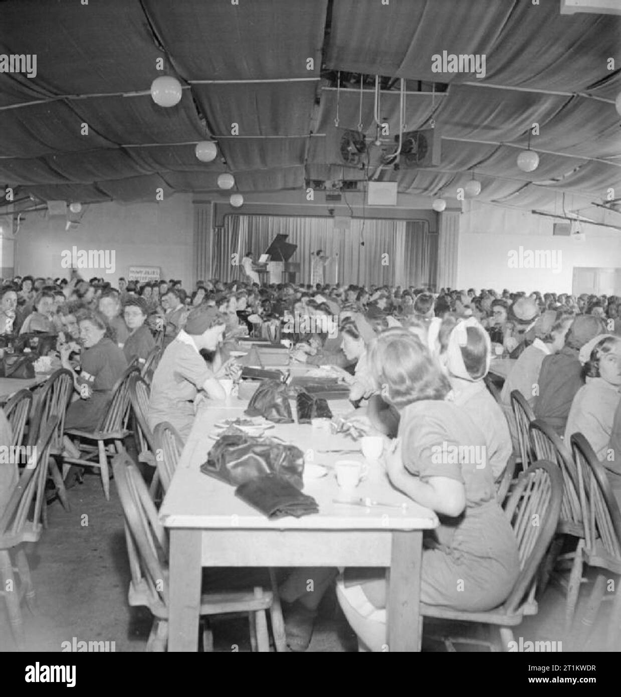 Welfare Facilities at a Royal Ordnance Factory, Britain, 1943 A wide view of war workers eating their lunch at the rows of long tables in the canteen of a Royal Ordnance Factory, somewhere in Britain. As they eat, they are listening to a violinist and her piano accompaniment, just visible on the stage at the far end of the canteen. A sign on the back wall (in the top left hand corner of the photograph) reads 'Radway Jollies Concert Party'. Stock Photo
