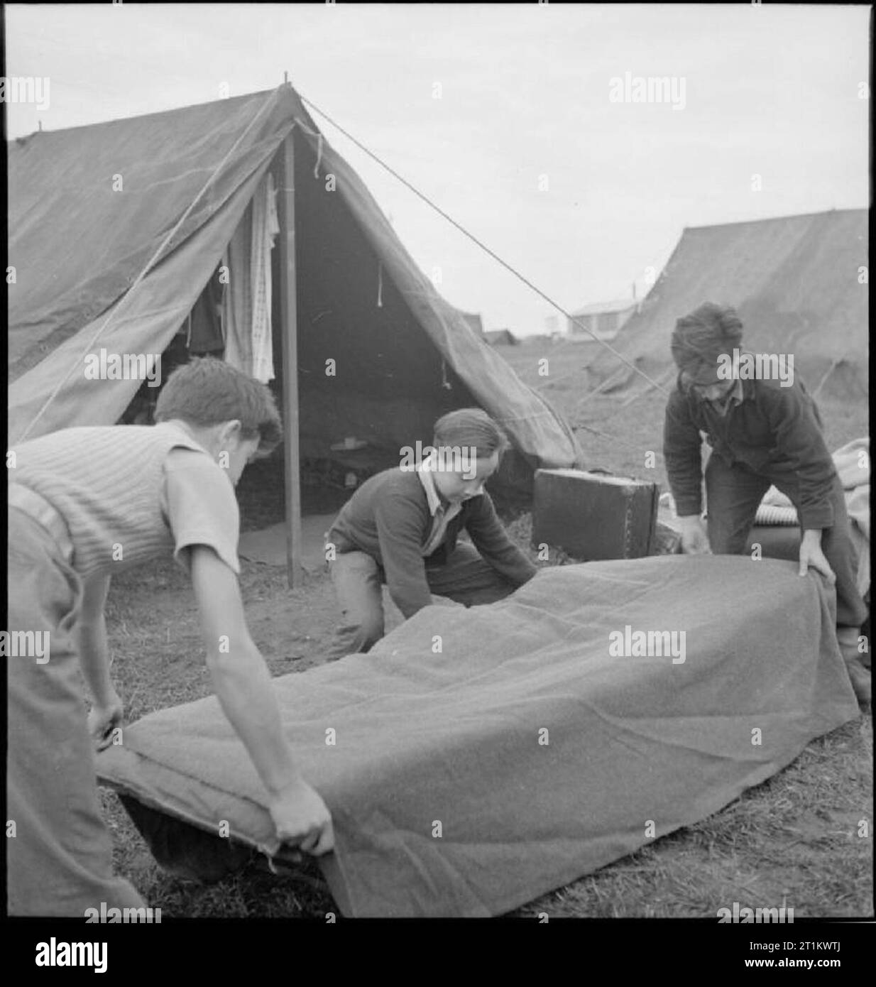 Youth Service Volunteers Help British Farmers- Agricultural Camp at Nunney Catch, Somerset, England, UK, 1943 Three boys make their beds at the Youth Service Volunteer camp at Nunney Catch, in between breakfast and work parade. Left to right, they are: Javier Sanchez (aged 16 1/2), Venanchio Zornova (aged 14) and his brother Mario Zornova (aged 16). All were refugees from the Spanish Civil War, and are on holiday from boarding school. The beds are being made outside the tents, which can be seen behind the boys as they work. Stock Photo