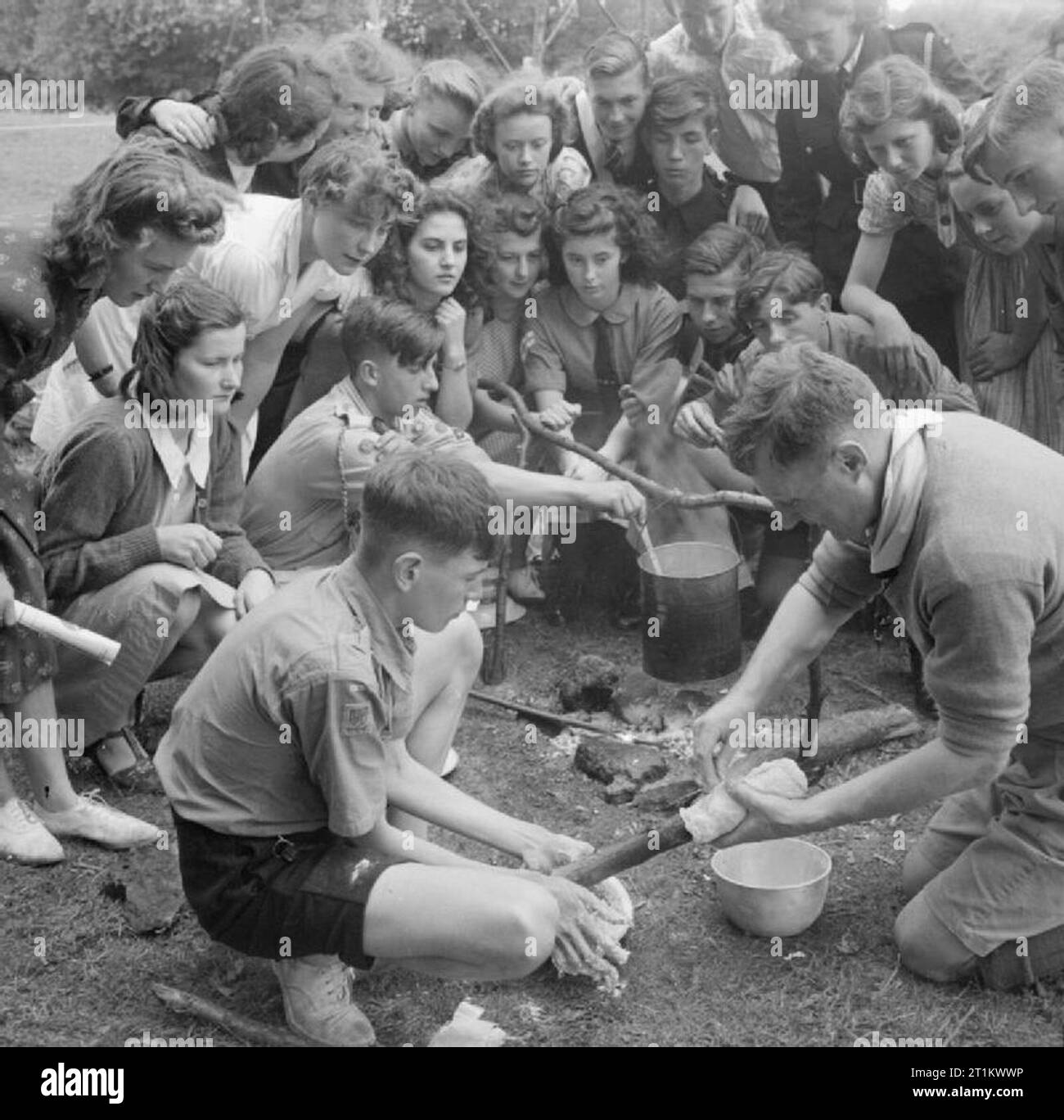 Youth Gathering- Residential Training For Youth Organisations, Sidcot School, Winscombe, Somerset, England, UK, 1943 Boy Scouts give a demonstration of camp bread making as part of the week-long youth training course being held at Sidcot School. Young people from other youth organisations have crowded around the Scout and his instructor and are watching the process intently. Another Scout can be seen stirring a billy can which is hanging over the camp fire. Stock Photo