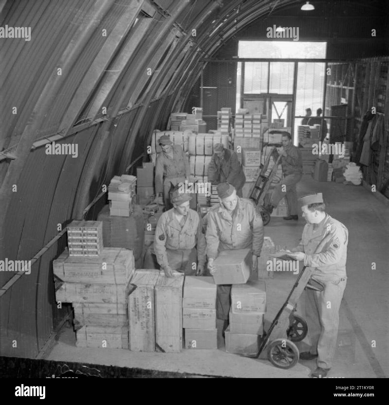 Britain Supplies US Army Store- Americans in Britain, 1943 GIs at work at an American Army store, somewhere in Britain. This particular area of the store supplies goods for Post Exchanges (also known as PXs), including candles, chocolate, shaving cream, toothpaste, batteries, ink, writing paper, shoe polish and playing cards. Making up a load for dispatch in the foreground are (left to right): T/5 Joe Brethauer (of Rote 2, Box 245, Fort Lupton, Colorado), Pfc Bernard Hanson (of 148 Magnolia Avenue, Kearny, New Jersey) and Staff Sergeant Robert McKenna of Big Bend, Wisconsin. Stock Photo