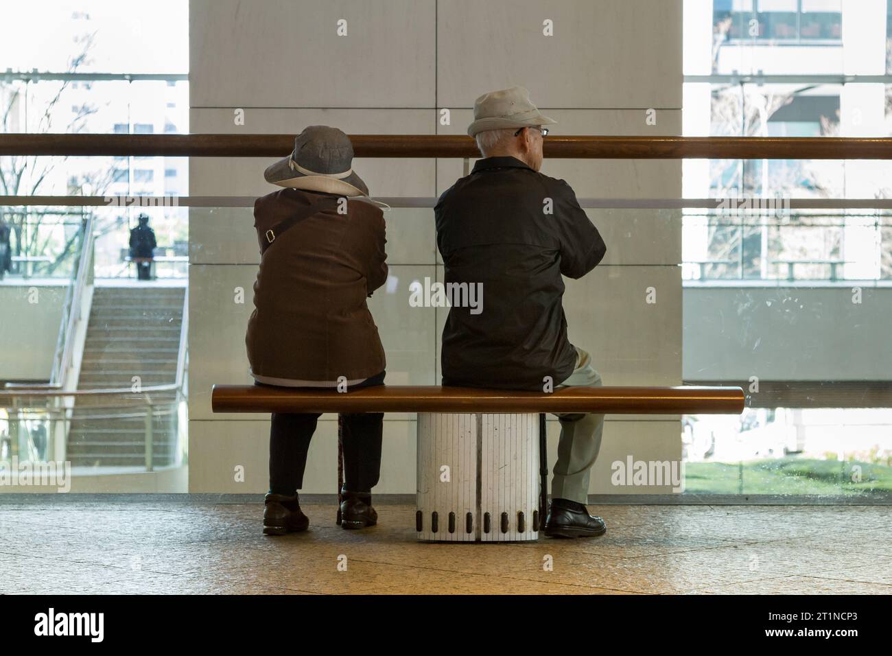 An older Japanese couple sit on a bench behind a pillar in a mall in Sakuragicho, Yokohama, Kanagawa, Japan Stock Photo