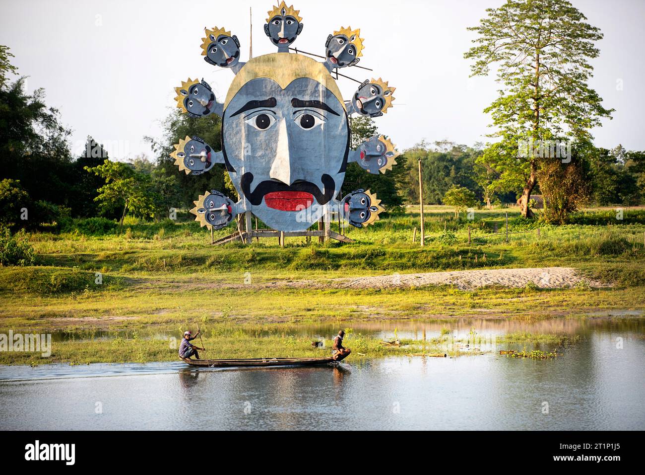 Fisherman paddling in a traditional wooden fishing boat on the river on Majuli island in Assam, passing giant mask on a river bank, India Stock Photo