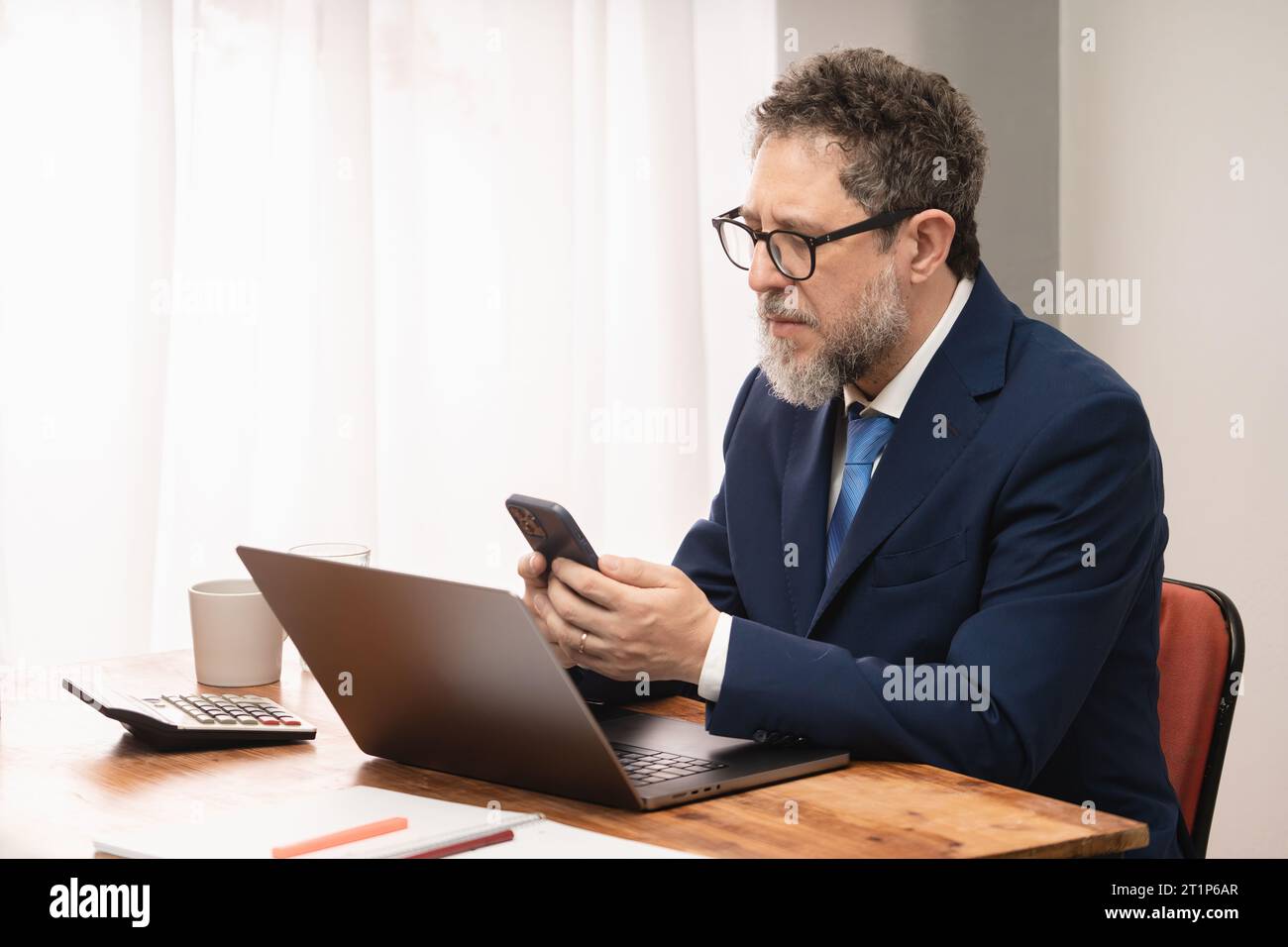 Mature businessman in blue blazer attentively checking messages on his smartphone in a well-lit office. Stock Photo