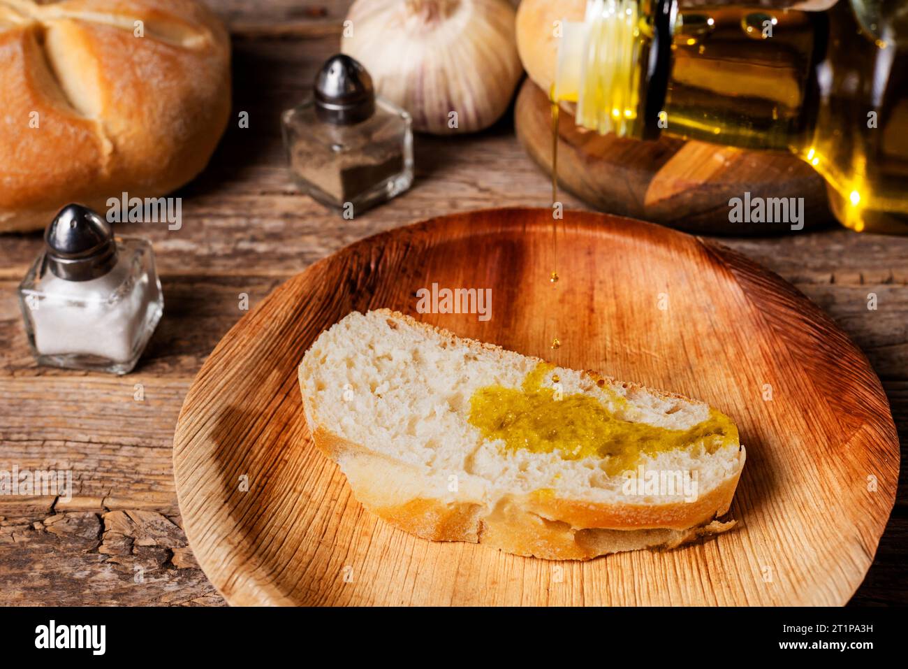 Bottle of olive oil pouring oil on a slice of bread, next to a shaker of salt, pepper, garlic and a piece of bread. Stock Photo