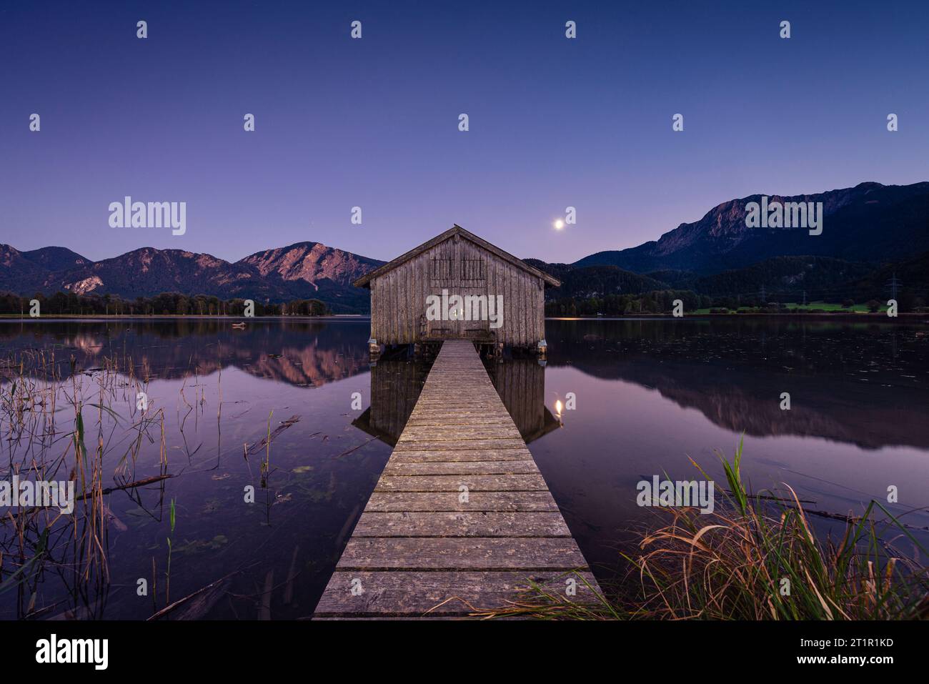 Moonrise over a wooden boathouse on the shore of the Kochelsee lake in front of the Herzogstand and Jochberg at dusk in autumn, Bavaria, Germany Stock Photo