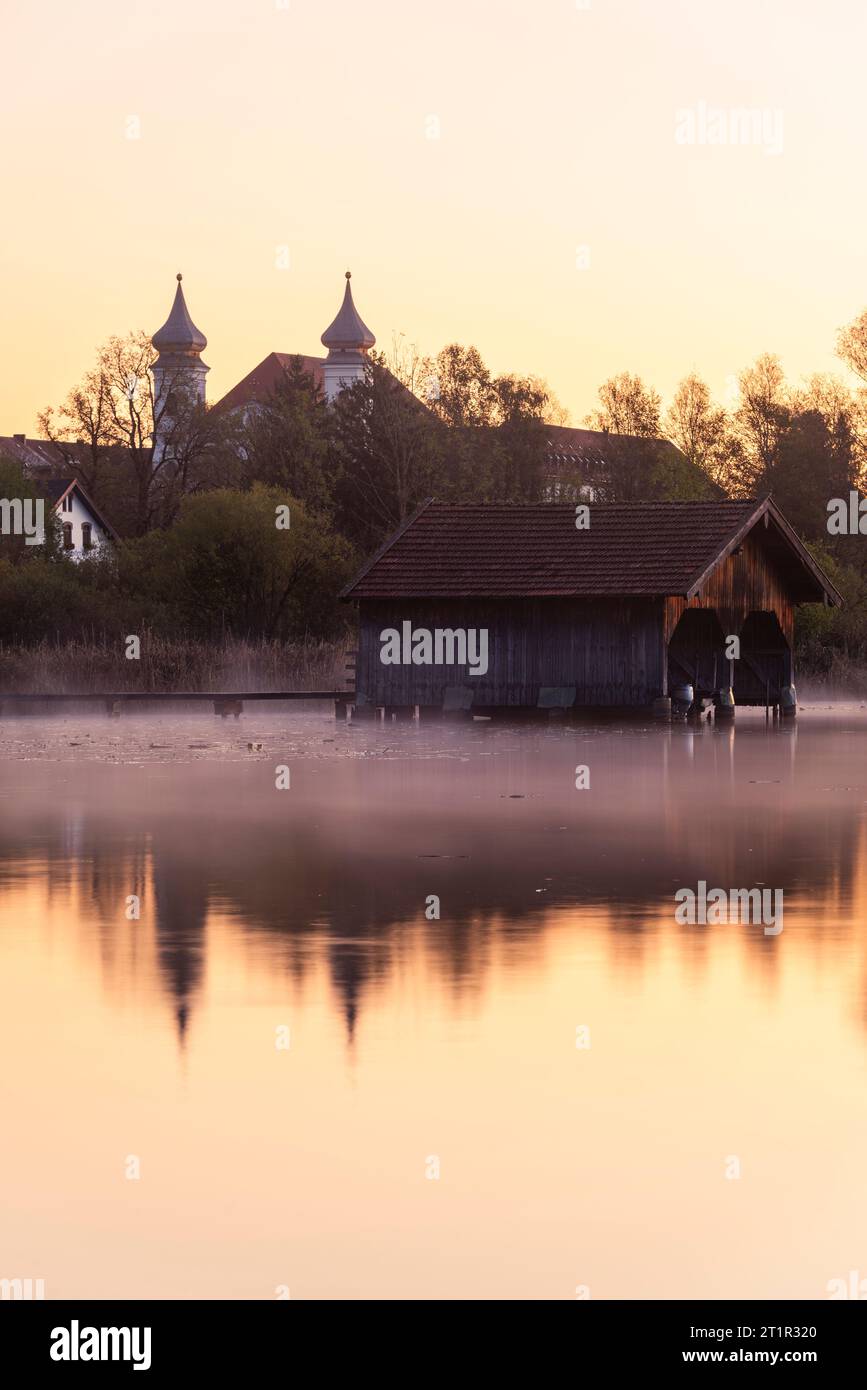 Boathouse in the fog on the shore of the Kochelsee lake in front of the panorama of the monastery in Schlehdorf in the golden dawn, Bavaria, Germany Stock Photo