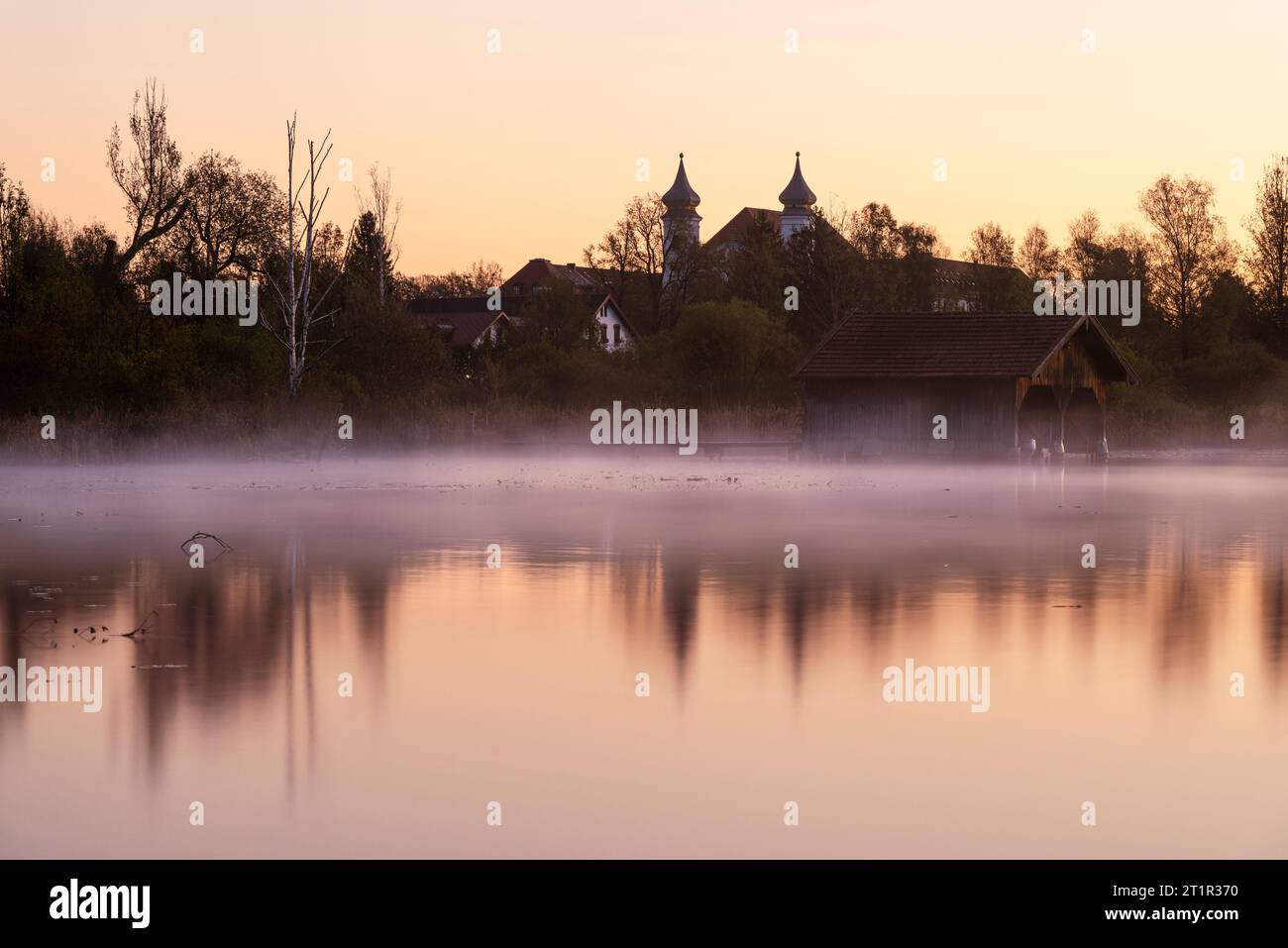 Boathouse in the fog on the shore of the Kochelsee lake in front of the panorama of the monastery in Schlehdorf in the golden dawn, Bavaria, Germany Stock Photo
