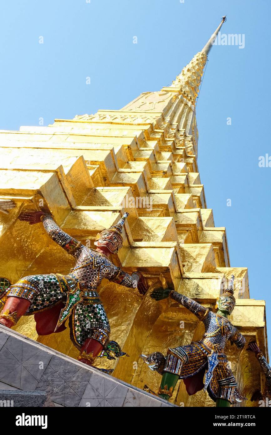 A golden temple spire in Thailand, supported by statues of mythical creatures decorated with colorful mosaic tiles, against a clear blue sky. Stock Photo