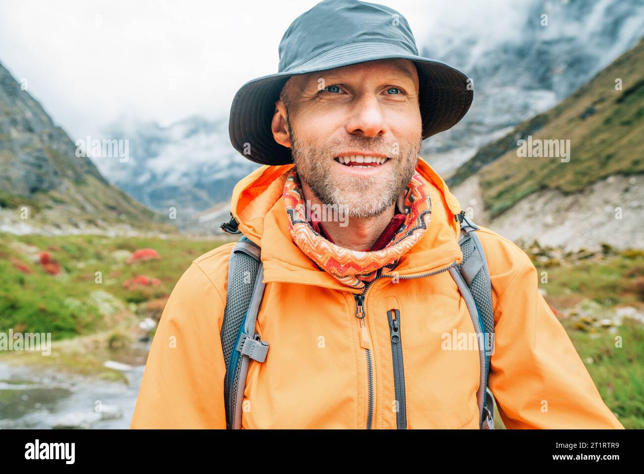 Portrait of smiling Man with backpack dressed orange waterproof jacket and funny hat walking the path during Makalu Barun National Park trek in Nepal. Stock Photo