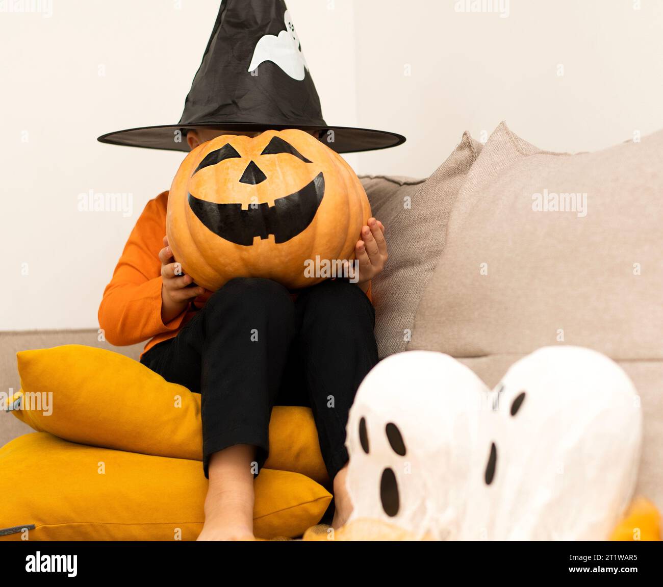 Halloween. Concept. A boy in an orange sweater and a haunted wizard's hat, with a pumpkin instead of a face, sits cheerfully in a home interior. Close Stock Photo