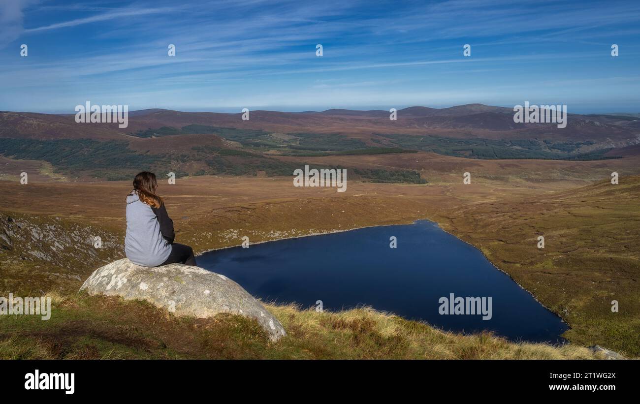 Young woman sitting on a rock and admiring a beautiful view of heart shaped lake, Lough Ouler and mountain range, Wicklow Mountains, Ireland Stock Photo