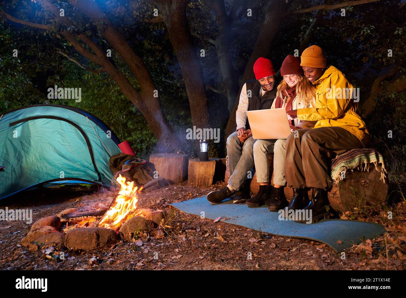 Three friends using laptop sitting together in front campfire one winter night. Happy young people. Stock Photo