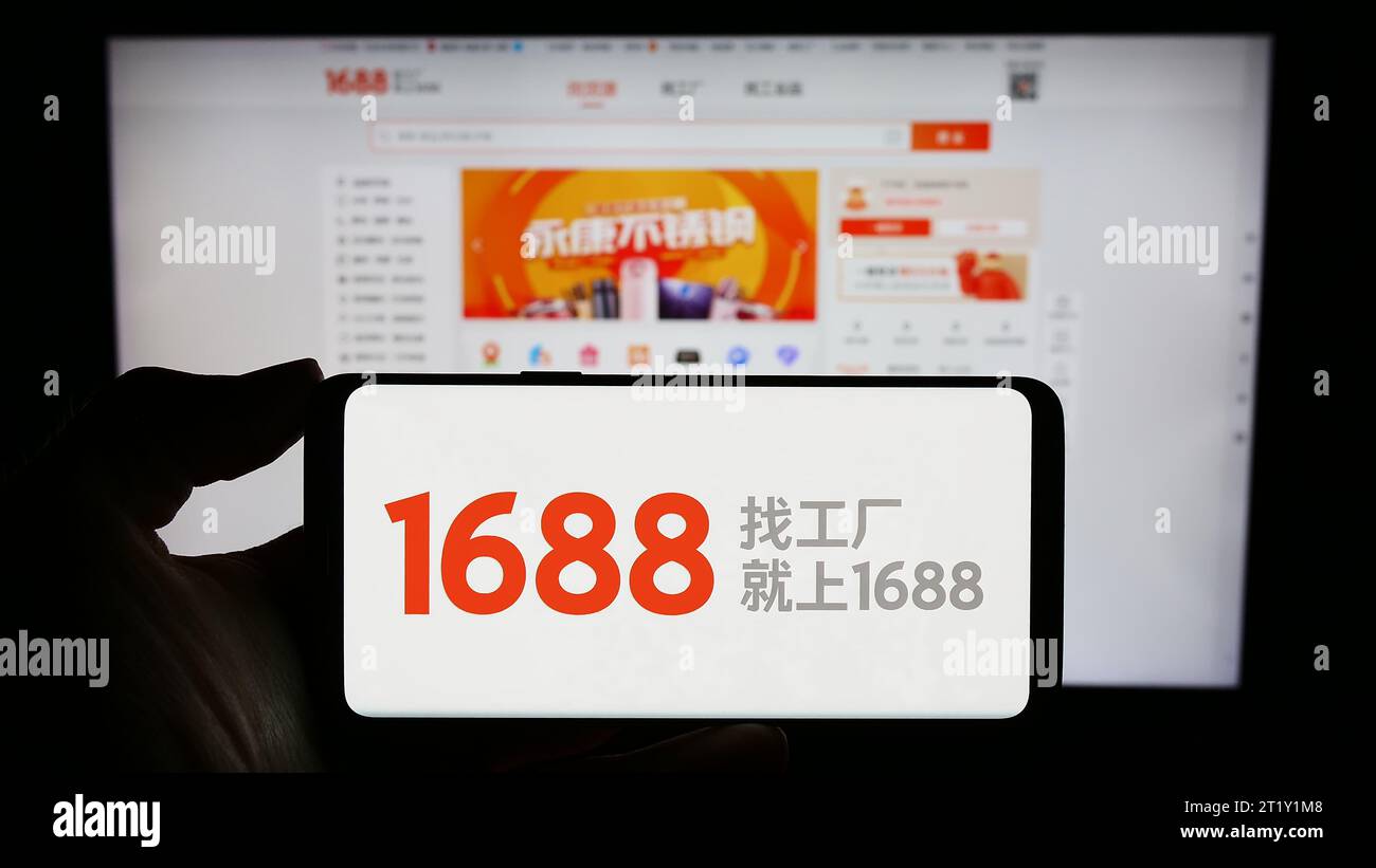 Person holding mobile phone with logo of Chinese online shop 1688.com (Alibaba) in front of business web page. Focus on phone display. Stock Photo