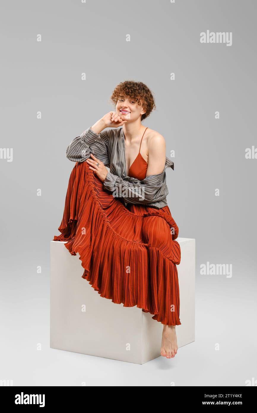 Smiling barefoot woman in sundress and shirt sits on cube in bright studio Stock Photo