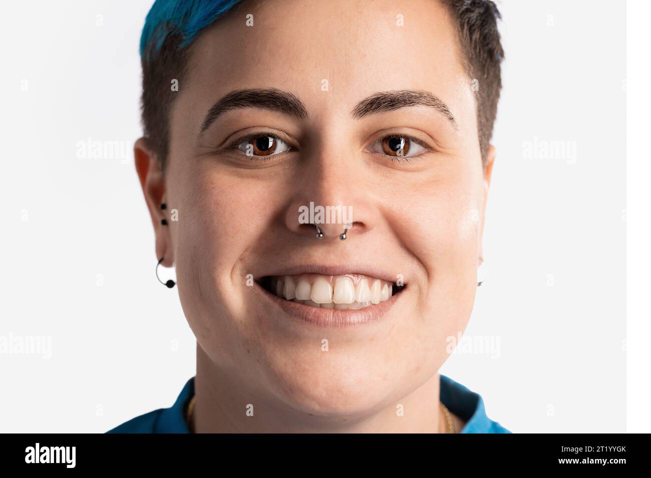 Close-up portrait of a smiling gender fluid woman, showcasing her androgynous features. She has vibrant blue short hair, a nose piercing, and an earri Stock Photo