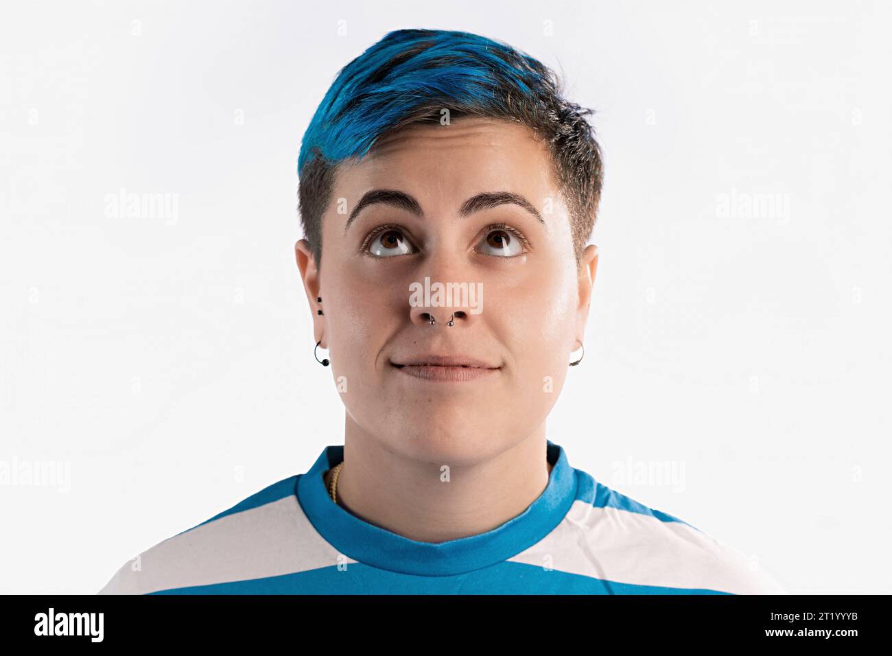 Half-length portrait of a gender fluid woman with vibrant blue short hair on a white background. She exudes a curious demeanor, looking upwards with a Stock Photo