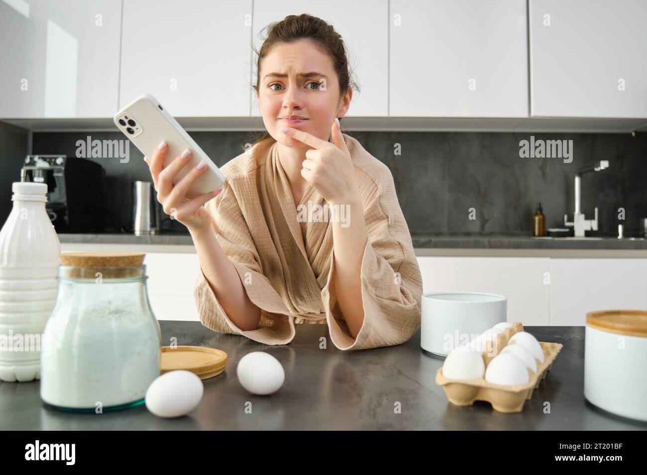 Attractive young cheerful girl baking at the kitchen, making dough, holding recipe book, having ideas. Stock Photo
