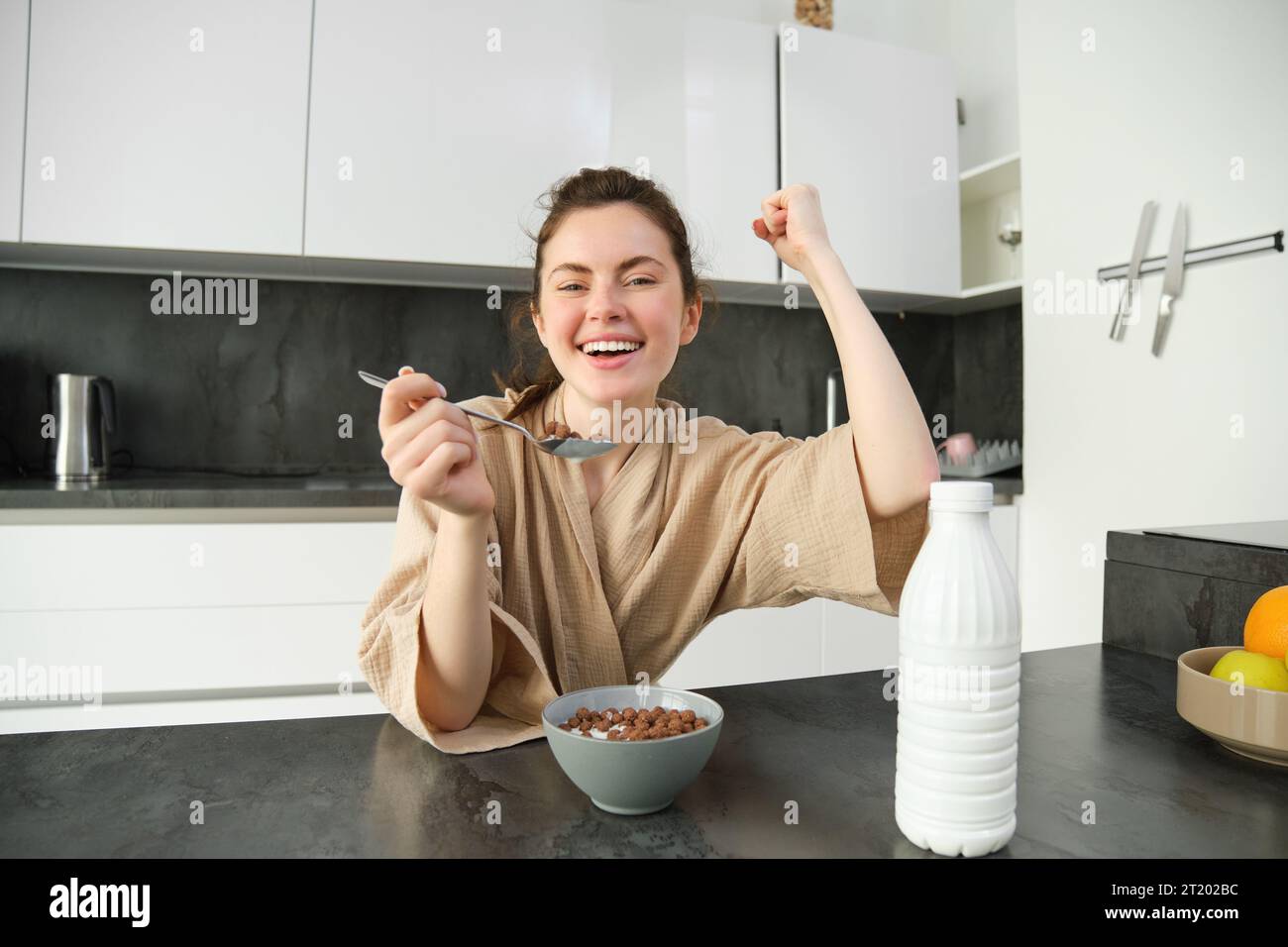 Portrait of enthusiastic young woman eating cereals with milk, looking excited and happy, sitting near kitchen worktop and having breakfast, raising h Stock Photo