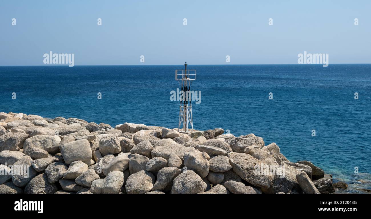 Metal beacon on rocky breakwater. Striped structure lighthouse, navigation sign on blue ripple sea background, sunny day. Stock Photo
