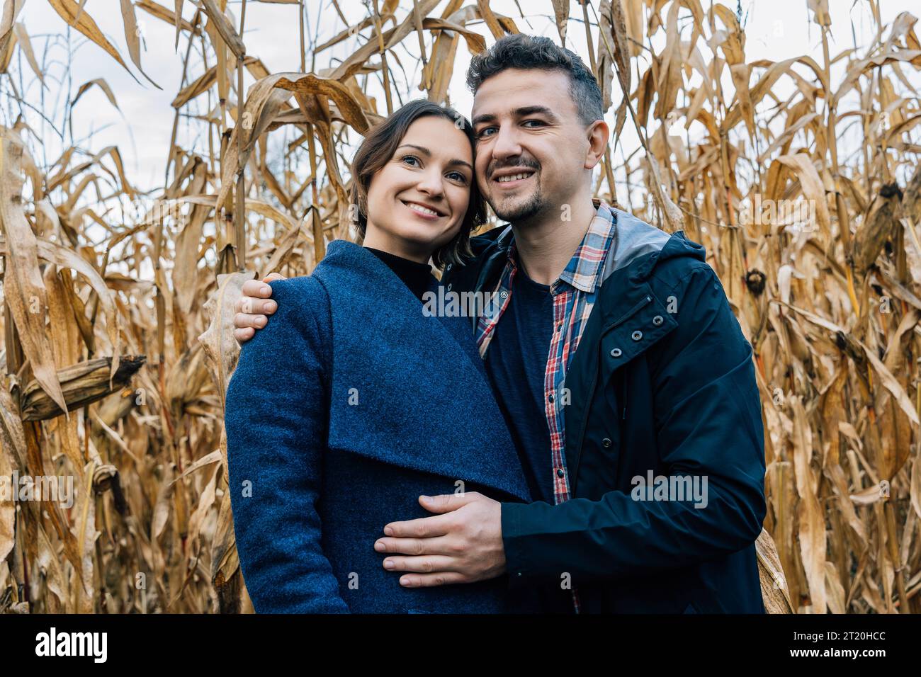 A happy couple in love stands in autumn near a cornfield. A man in a shirt and jacket hugs a woman in a coat Stock Photo