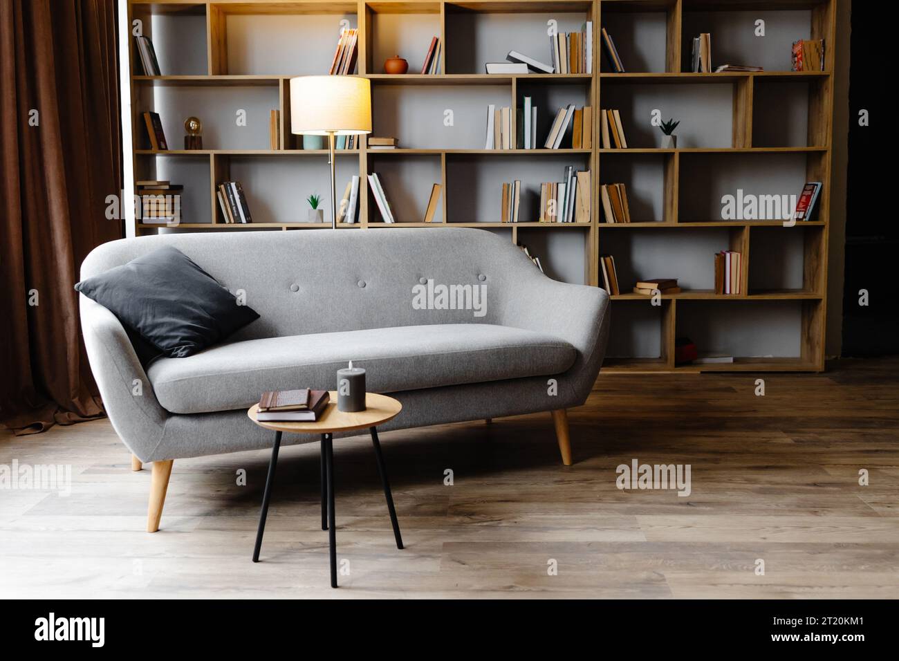 Grey sofa with cushions standing on living room with stylish interior design and collections books on bookshelves in library. Work cabinet in modern a Stock Photo