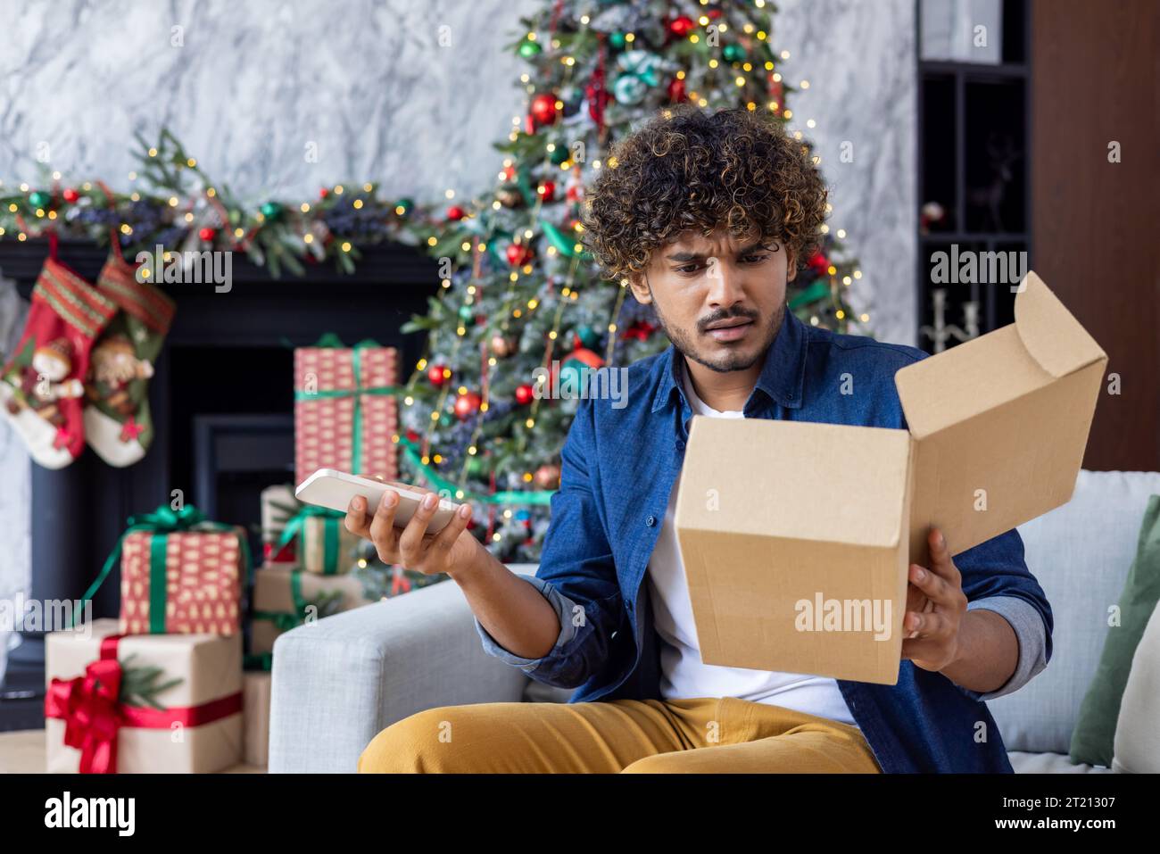 Man upset and disappointed received wrong spoiled package gift, Hispanic man for Christmas sitting in the living room, near the Christmas tree, holding the phone and calling customer service. Stock Photo