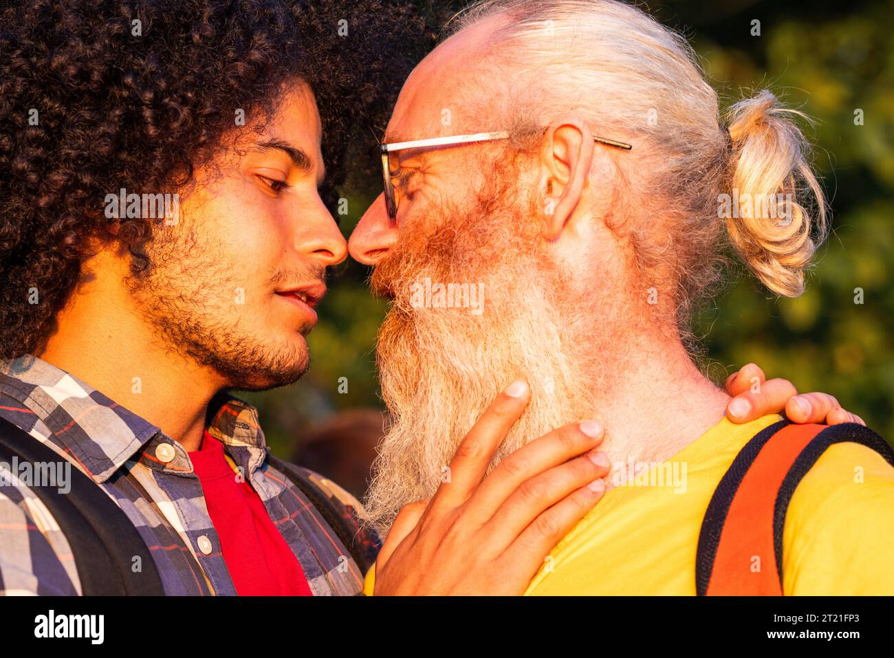 A tender and passionate moment captured as an attractive gay millennial Arabian man with long curly hair and his older Caucasian male lover with long grey hair and a beard gaze at each other, about to share a passionate kiss in the warm embrace of the setting sun. Intimate Sunset Moment, Gay Couple's Kiss. High quality photo Stock Photo