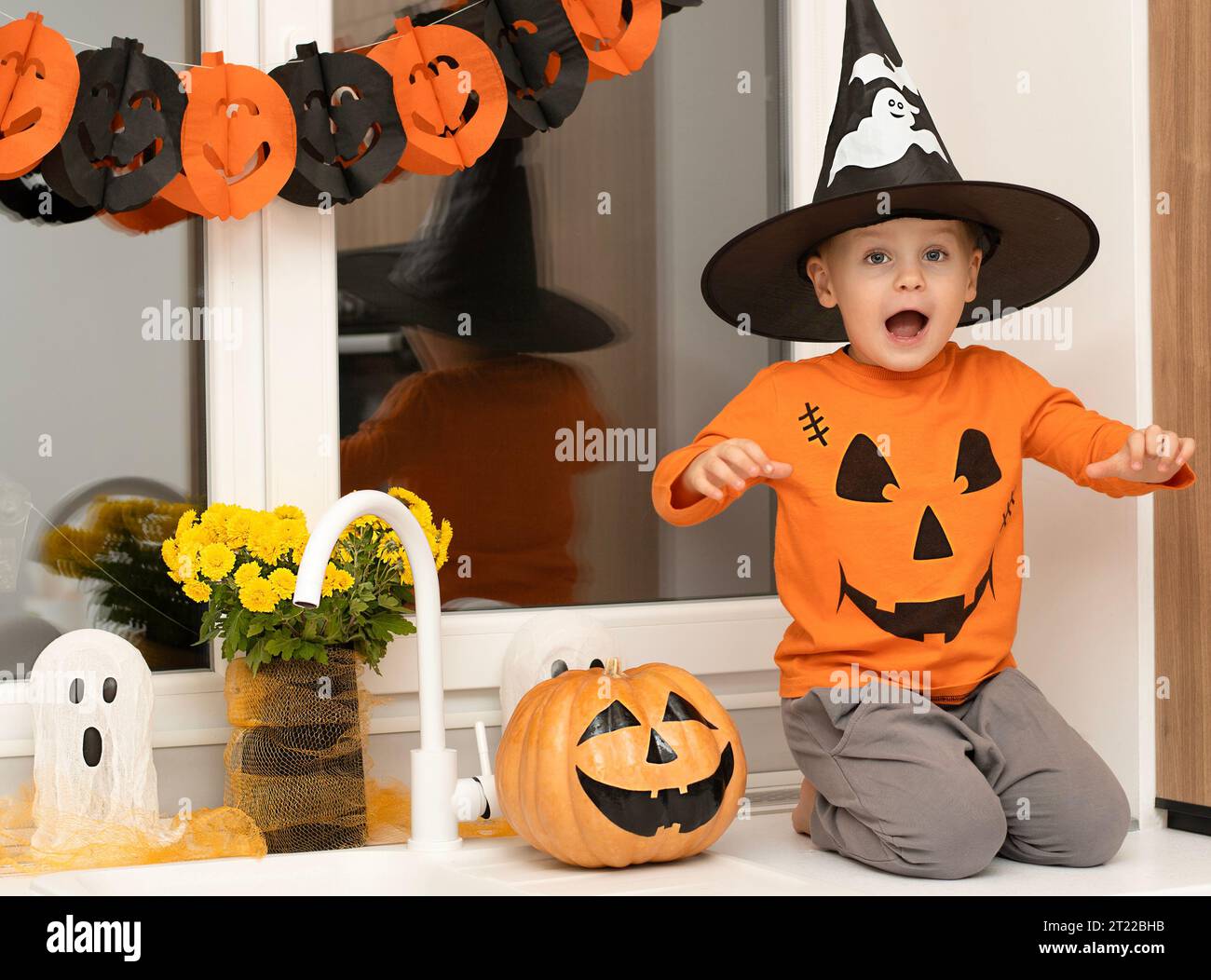 Halloween concept. A small, cheerful, handsome boy in a wizard's hat and an orange sweater sits on a table in the kitchen against the background of a Stock Photo