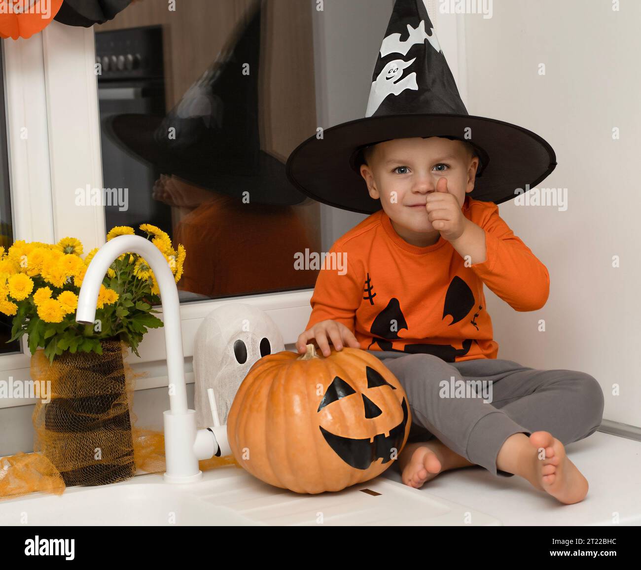 Halloween concept. A small, cheerful, handsome boy in a wizard's hat and an orange sweater sits on a table in the kitchen against the background of a Stock Photo