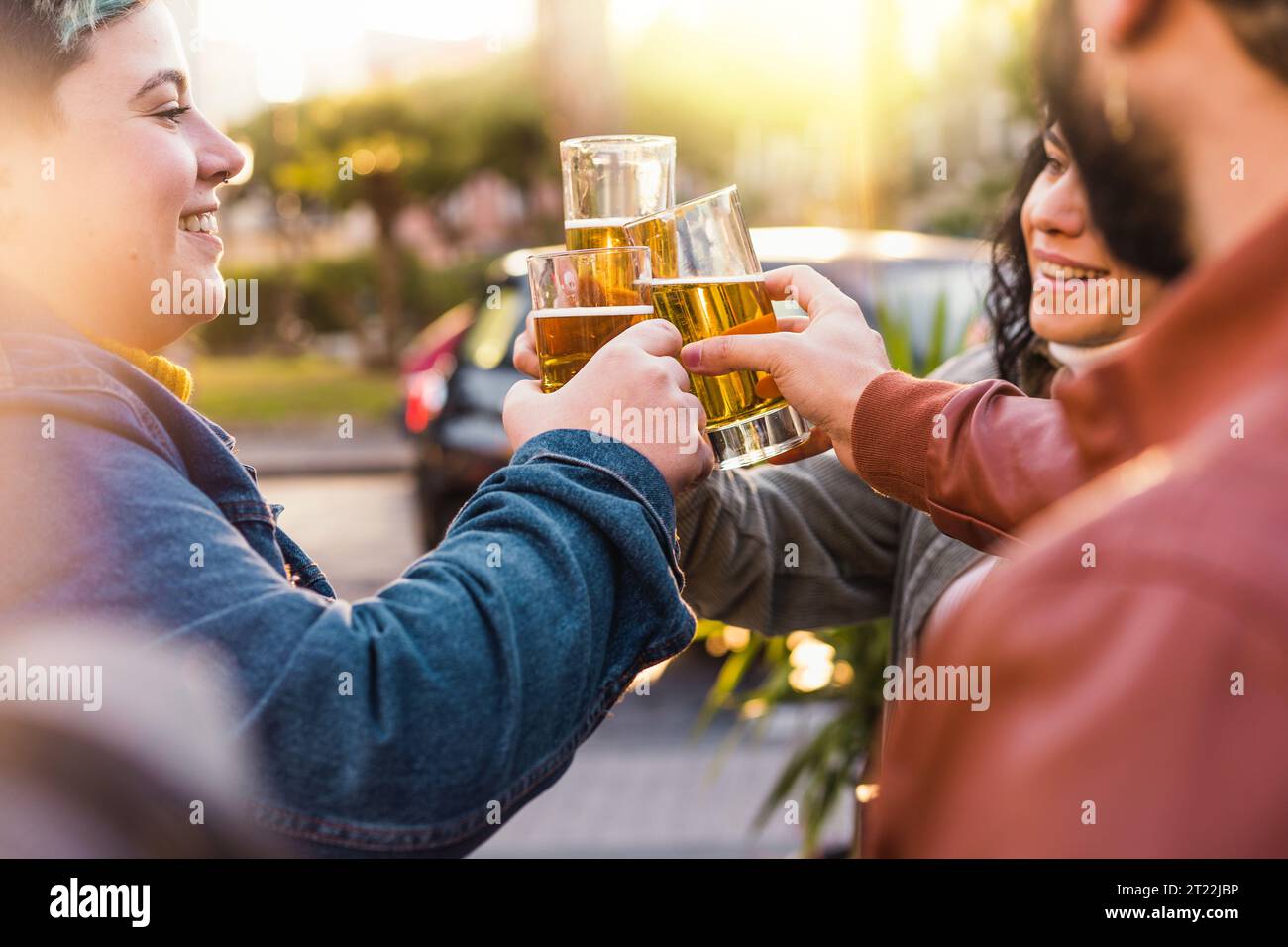 A diverse group of friends, including a gender fluid woman with vibrant short colored hair, raise their beer glasses in a toast at an outdoor sidewalk Stock Photo