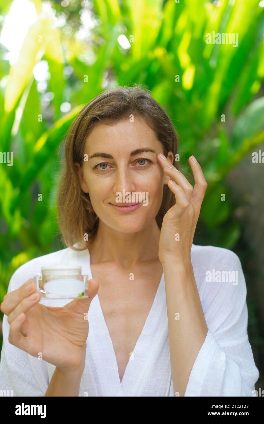 Vertical close up photo of the face of a middle-aged woman. A woman applies cream under her eyes with massage movements and holding a jar in her hand. Stock Photo