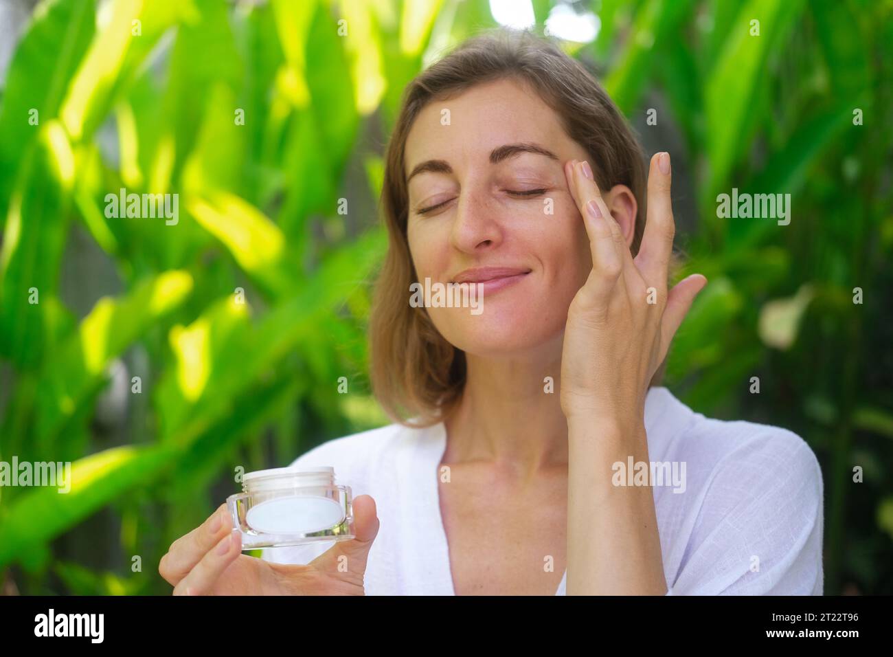 Close up photo of the face of a middle-aged woman. A woman applies cream under her eyes with massage movements and holding a jar in her hand. Anti agi Stock Photo