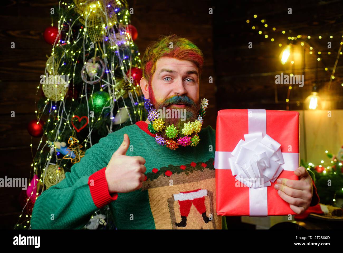 Smiling bearded man with decorated Christmas beard hold Christmas present shows thumb up. Christmas beard decorations. Delivery service. Happy New Stock Photo