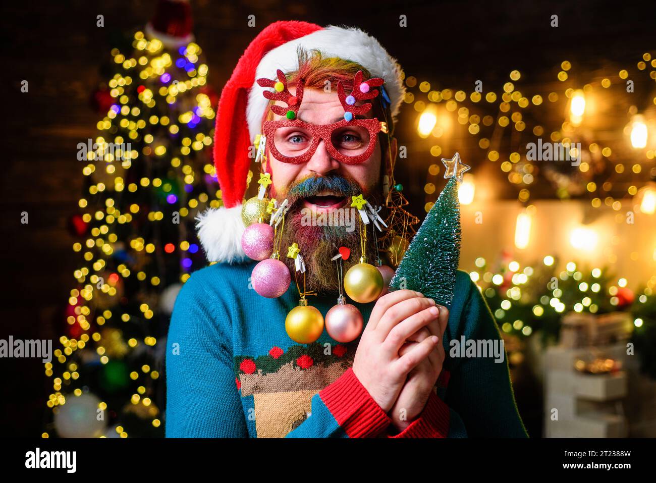 Merry Christmas and Happy New year. Smiling man in Santa hat with decorated beard holds small Christmas tree. Christmas beard style. Bearded Santa Stock Photo