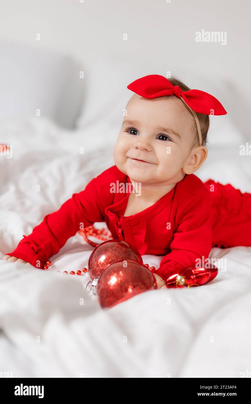 Cute happy baby in a red jumpsuit is lying on her stomach in bed Stock Photo