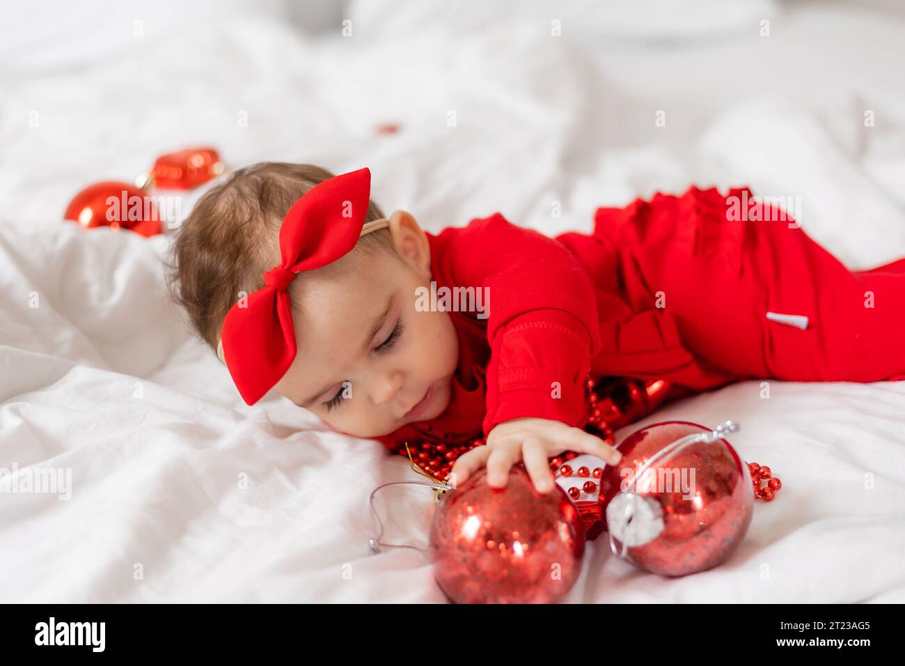 Cute happy baby in a red jumpsuit is lying on her stomach in bed Stock Photo