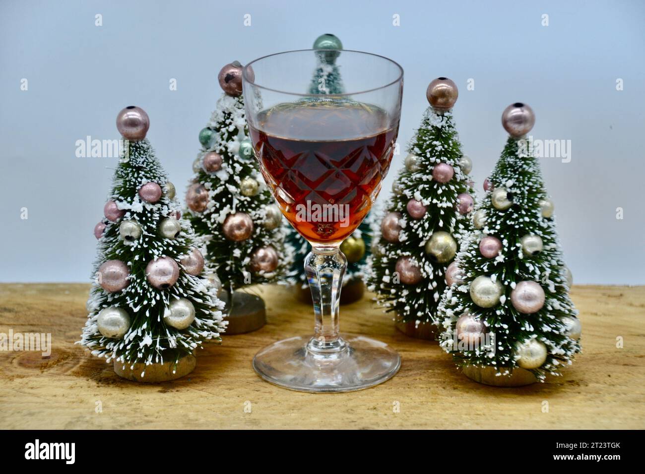 A single glass of sherry or port wine on a rough wooden table with five cute tiny snowy fake Christmas trees with colored bauble decorations close up Stock Photo