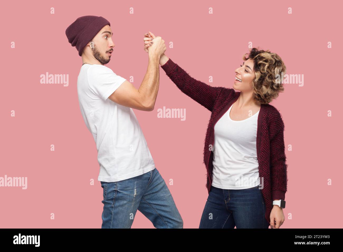 Portrait of smiling positive woman and surprised shocked man standing together and holding hands, looking at each other with different emotions. Indoor studio shot isolated on pink background. Stock Photo